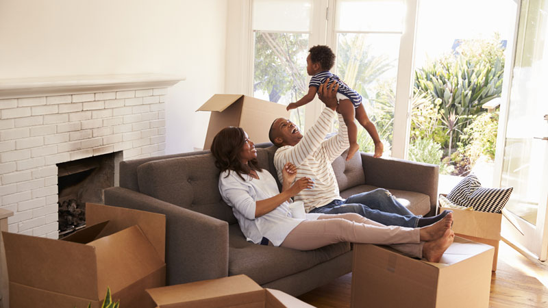 happy smiling family of father and mother lounging at the couch with moving boxes on the ground lifting toddler baby over father's head