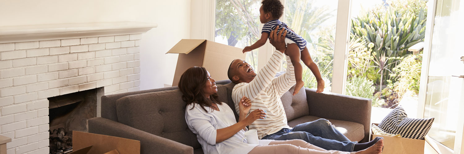 happy smiling family of father and mother lounging at the couch lifting toddler baby over father's head