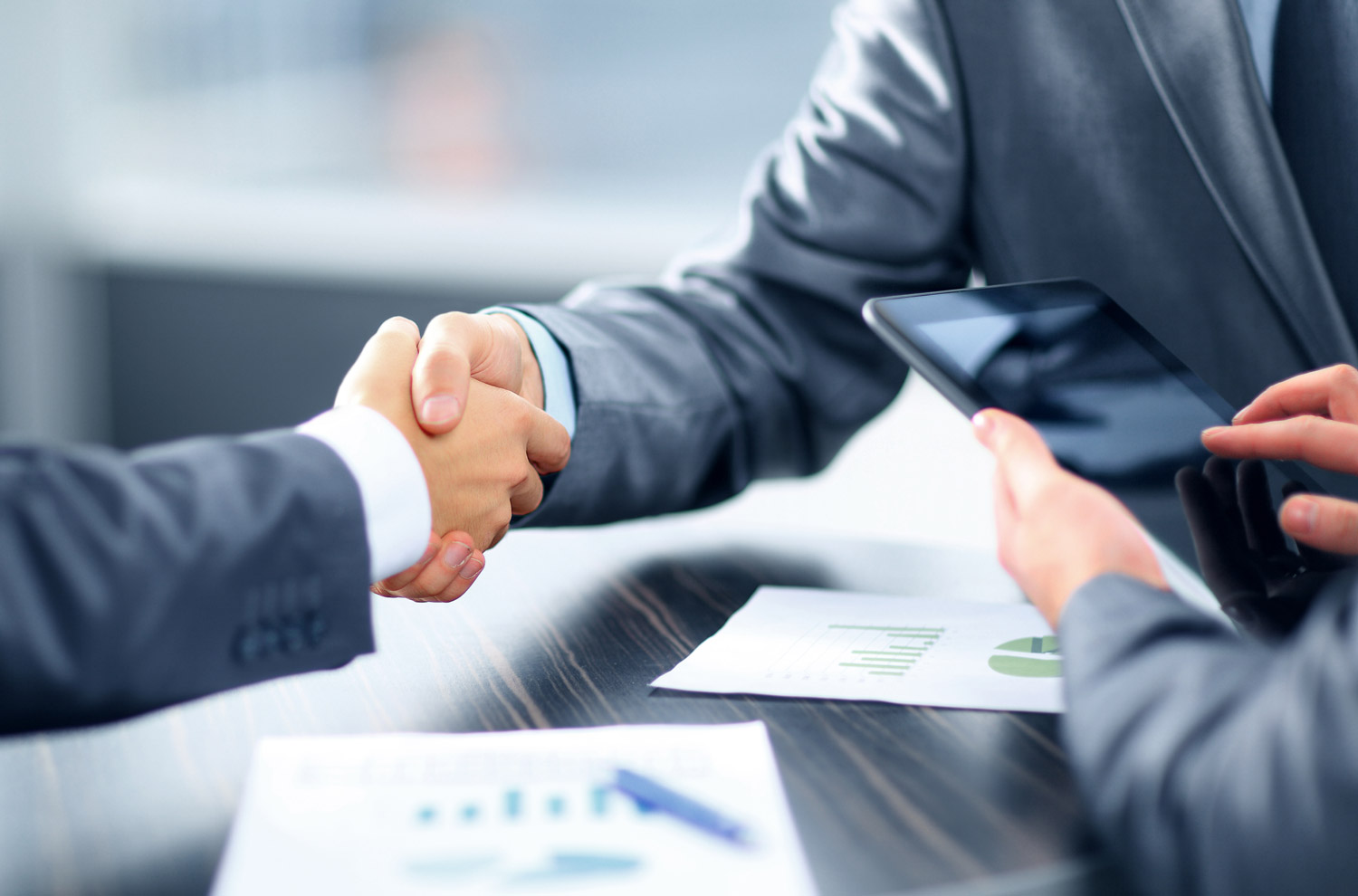 two professionals in grey suits shaking hands over the documents with graphical charts and another person using tablet